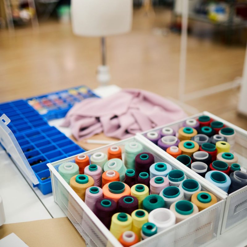 Workplace of tailor: close-up of two containers full of colorful spools of threads on table with mess in atelier